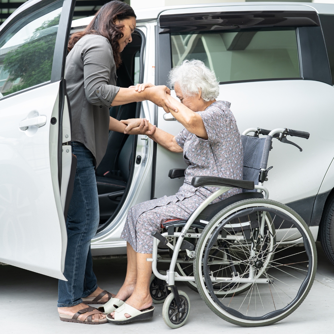 woman assisting a senior woman out of her wheel chair and into a vehicle